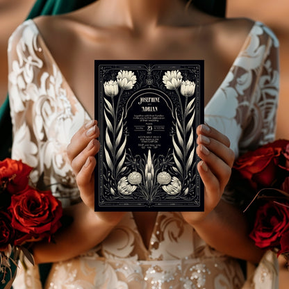 A close-up of a person holding a Gothic invitation in front of their wedding dress. The red rose bouquet in the background adds a bold, romantic touch to the vintage, moody setting. The names and event details are centered.